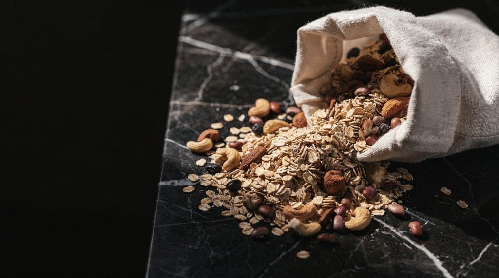 Rolled oats with almonds, cashews and beans spilling from a linen bag onto black marble